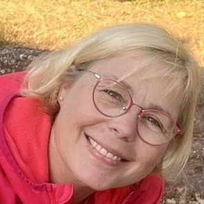 Geneviève Femme souriante avec des lunettes roses, portant un pull rouge, posé sur un sol en terre.